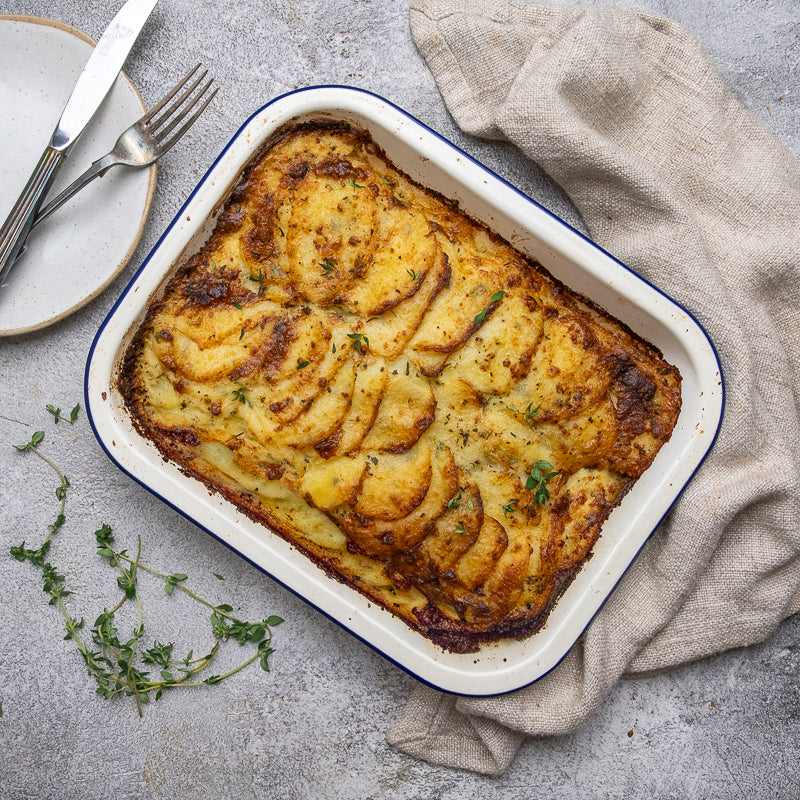 A baked potato casserole with sliced potato layers on top, served in a white rectangular baking dish with a blue rim. The dish is photographed from above on a gray stone-like surface with a beige cloth, a small plate with a knife and fork, and a few herb sprigs nearby.