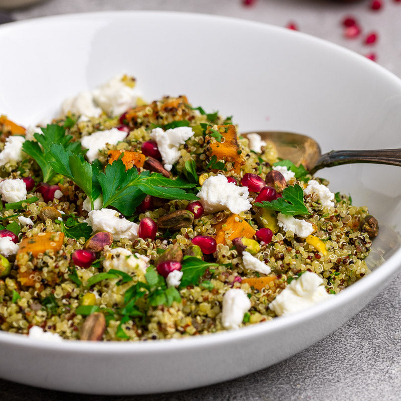 A bowl of quinoa salad topped with pieces of goat cheese, pistachios, and pomegranate seeds, alongside herbs and a pomegranate fruit in the background.