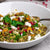 A bowl of quinoa salad topped with pieces of goat cheese, pistachios, and pomegranate seeds, alongside herbs and a pomegranate fruit in the background.