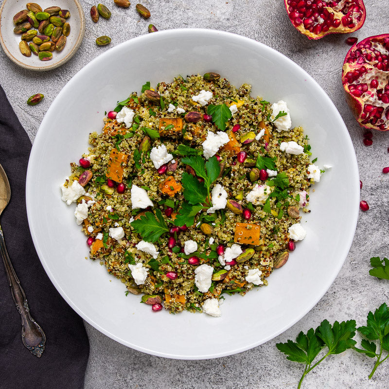 A bowl of quinoa salad topped with pieces of goat cheese, pistachios, and pomegranate seeds, alongside herbs and a pomegranate fruit in the background.