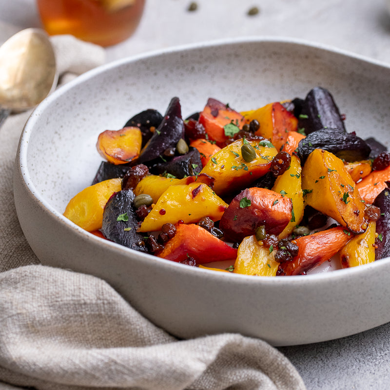 A close-up of a ceramic bowl filled with roasted colorful vegetables, including yellow squash, orange carrots, and dark purple beets, garnished with herbs and capers, set on a neutral textured surface with a linen napkin.