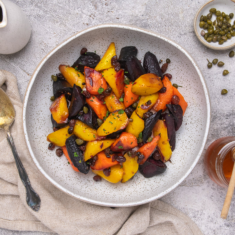 Overhead shot of a speckled white bowl filled with roasted sweet potatoes, carrots, and beets with capers and herbs, set on a textured gray surface with a spoon, napkin, and small garnish bowls.