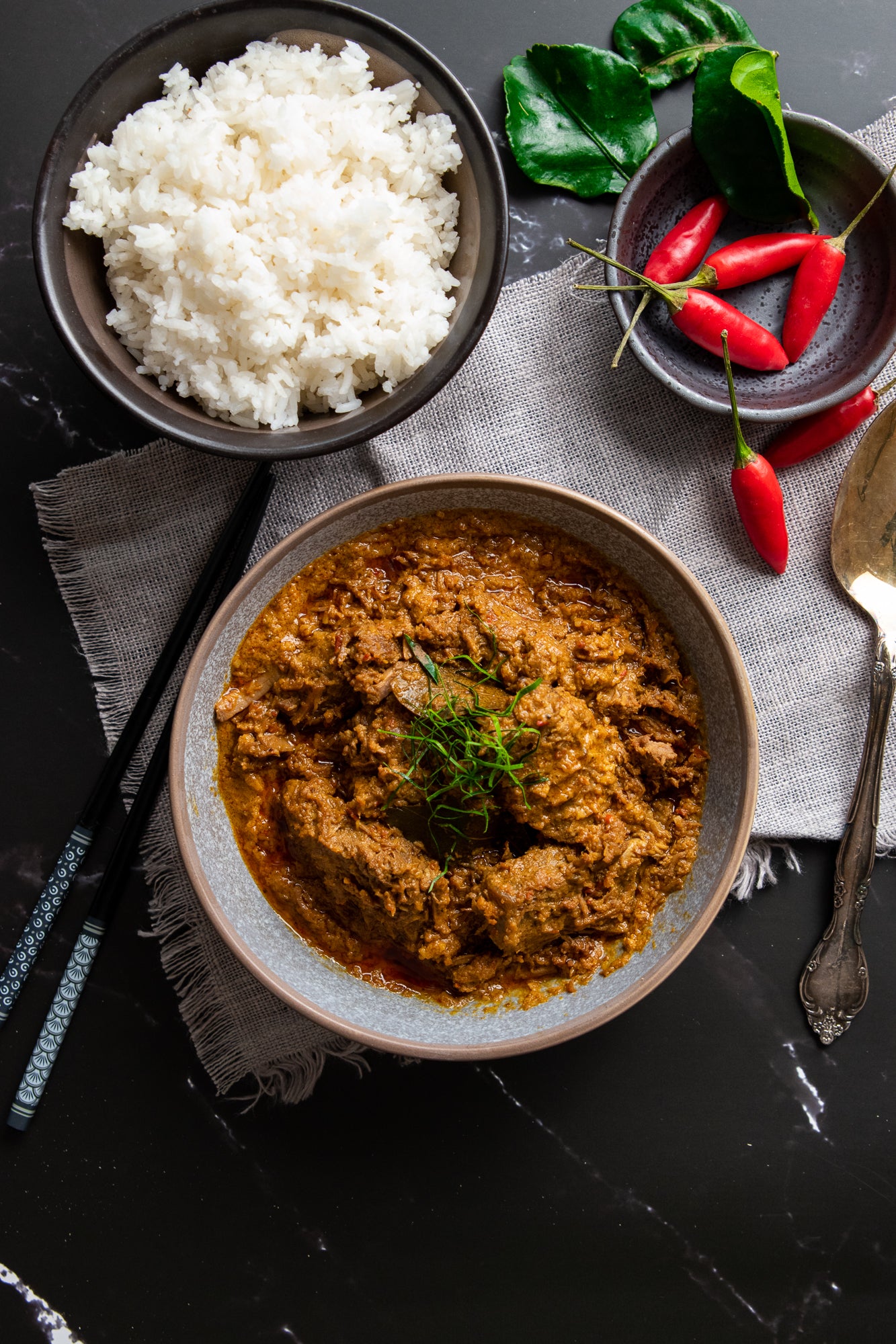 Bowl of rendang with a side of rice and red peppers on a dark surface
