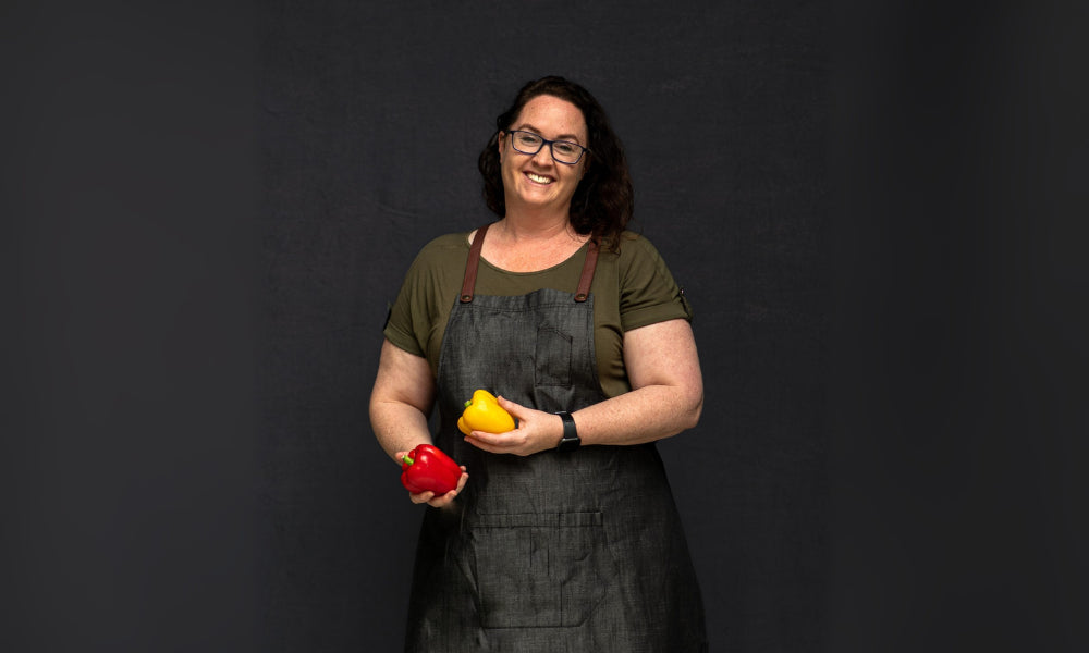 Bec, FoodSt cook wearing a dark apron holding red and yellow bell peppers against a black background