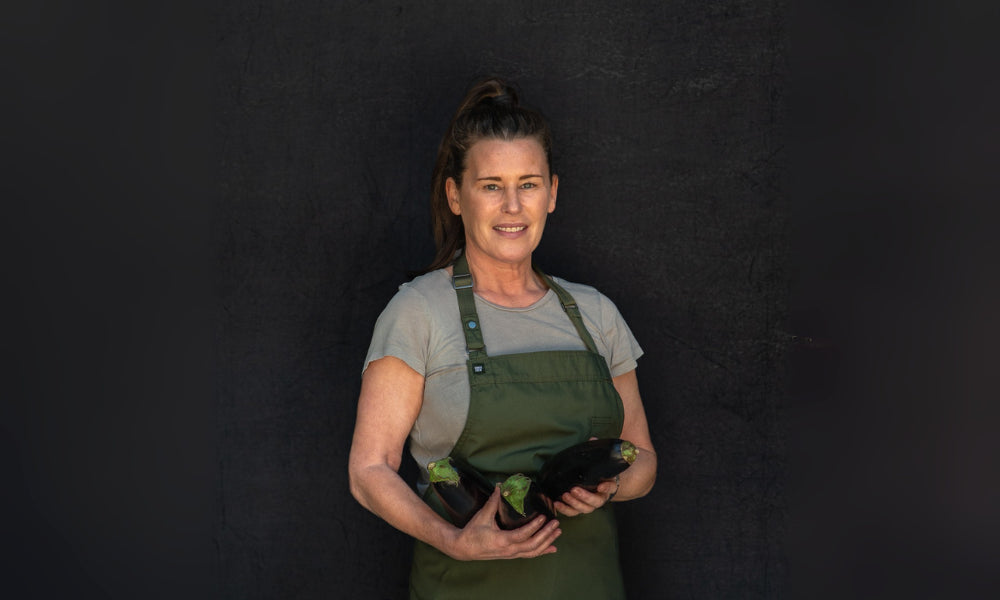 Image of FoodSt cook Denise wearing a green apron holding black pots against a dark background