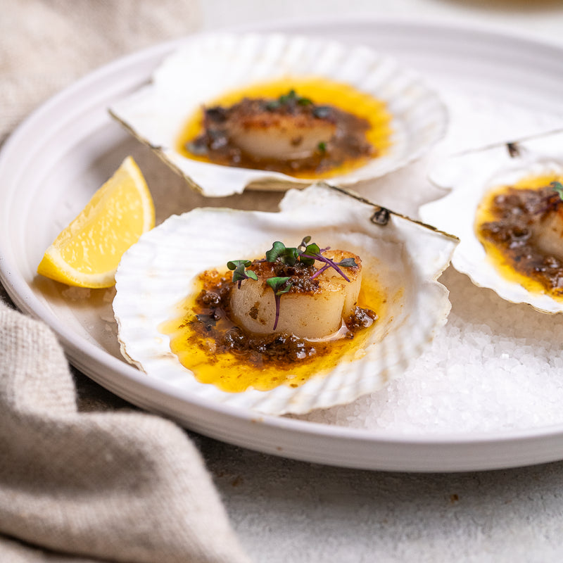 Three cooked scallop shells on a white plate with crushed ice, a lemon wedge, and a beige cloth. The scallops are topped with a glossy orange sauce, crispy fried bits, and green herb garnish.