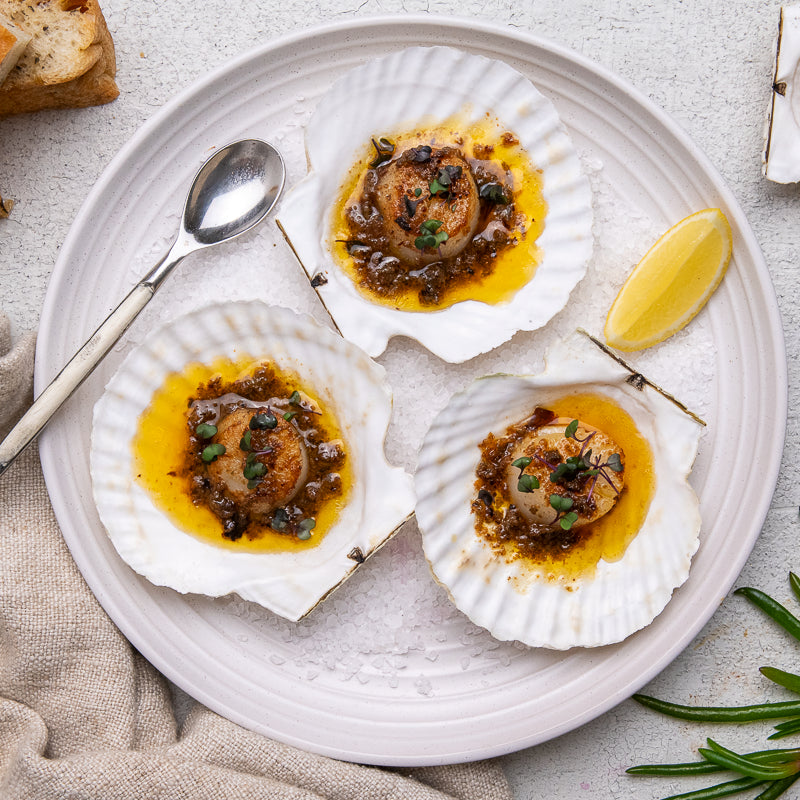 Three cooked scallop shells on a white plate, topped with golden oil, a dark sauce, and chopped green herbs, with a lemon wedge and spoon on a light textured surface.