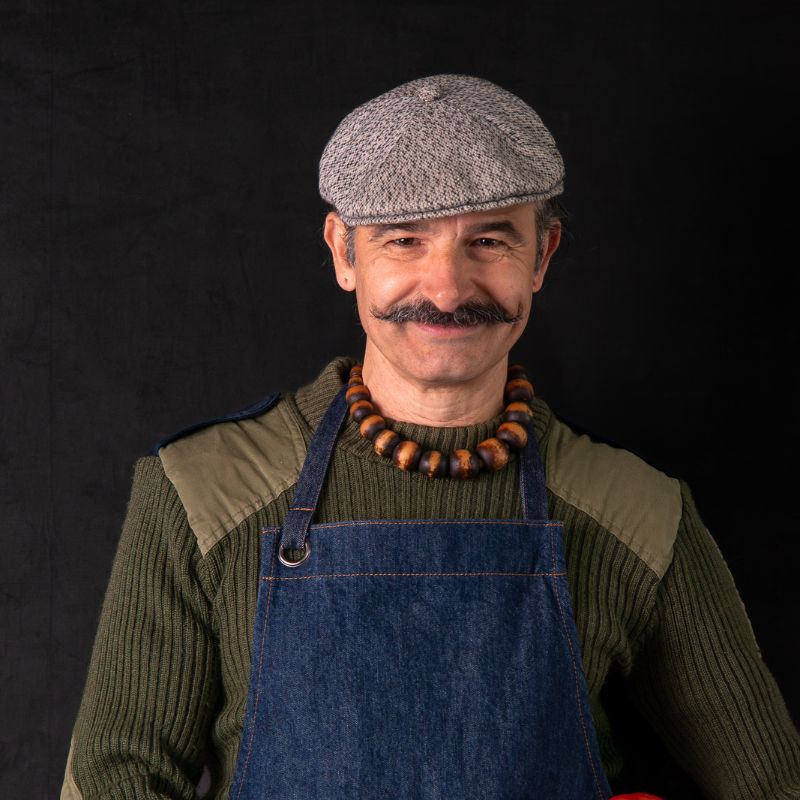 Image of FoodSt cook Simone wearing a flat cap, sweater, and apron against a black background