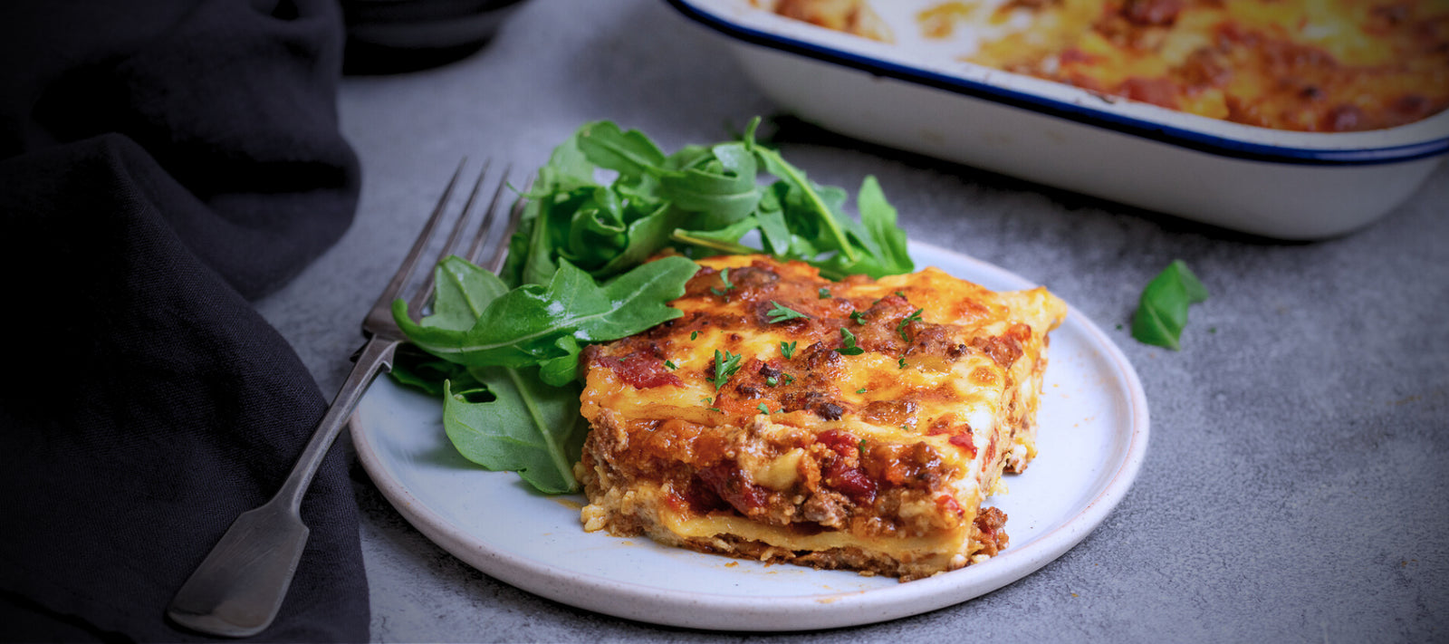 Slice of Davide's lasagna on a white plate with a side of greens, placed on a gray surface.