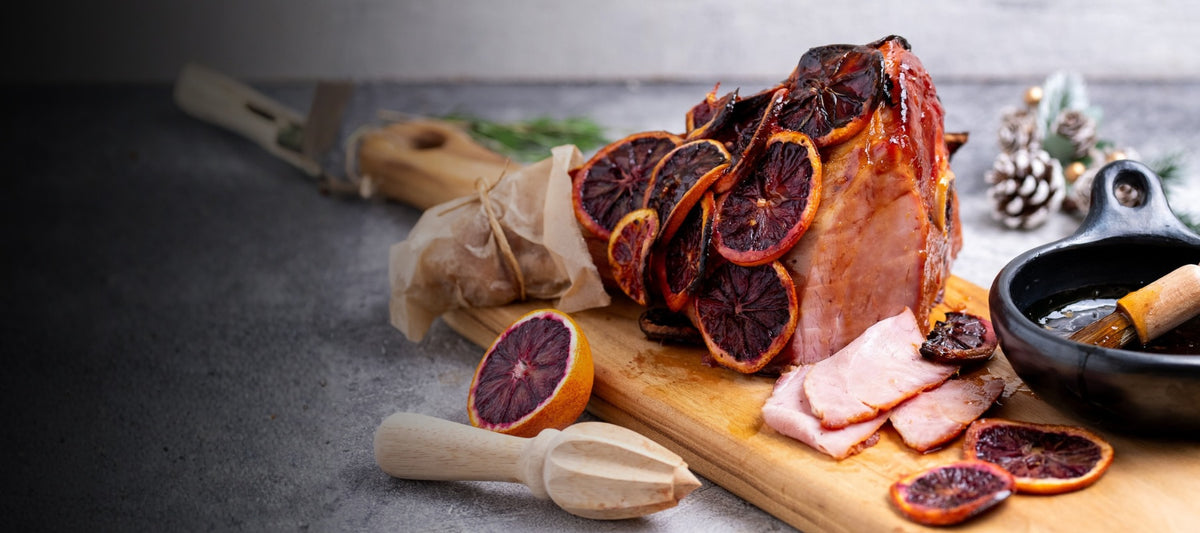 A styled food scene showing a sliced ham or cured meat on a wooden cutting board, topped and surrounded by dried blood orange slices, with a small bowl and utensils on a dark gray surface.