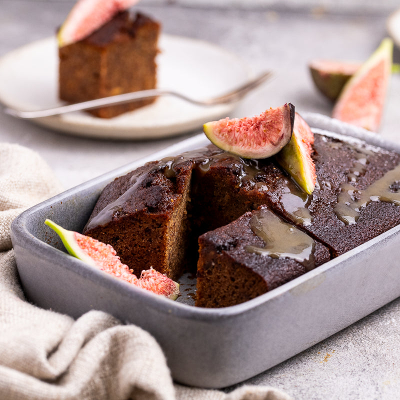 A rectangular gray baking dish holding a sliced chocolate cake or bread pudding topped with glossy syrup and fresh fig slices, set on a light surface with a folded beige cloth. A second slice sits on a plate in the blurred background.