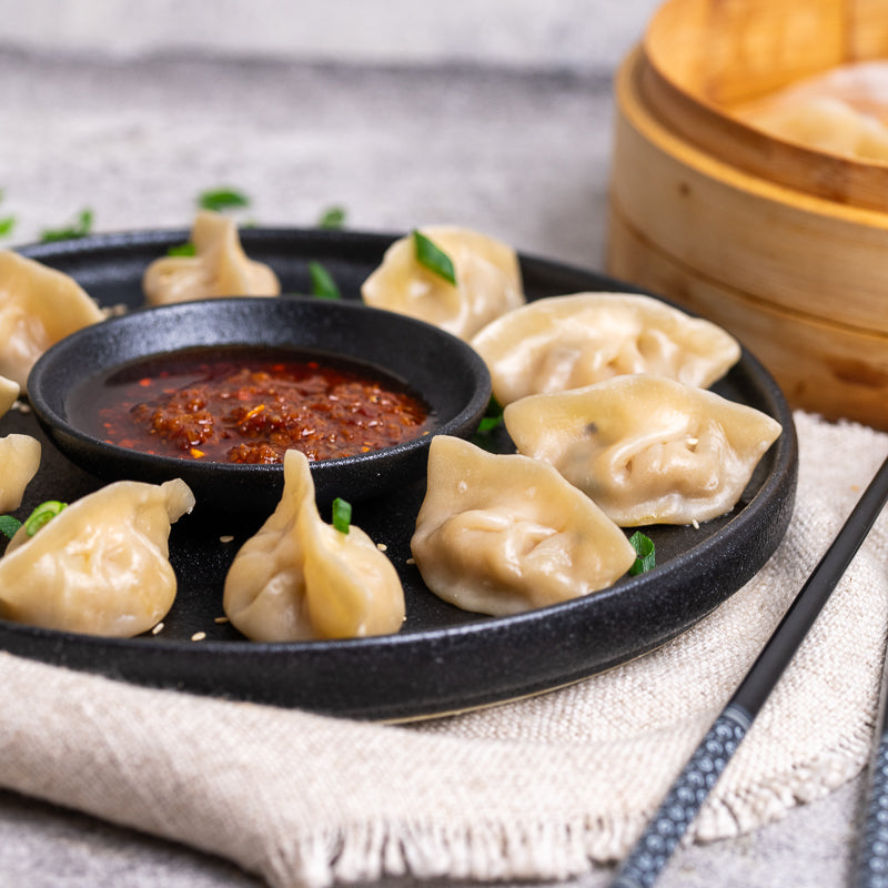 Dumplings on a black plate with a small bowl of red sauce, placed on a textured surface.