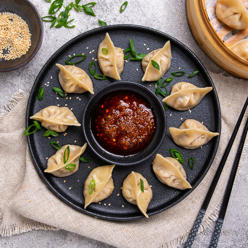 Dumplings on a black plate with a small bowl of red sauce, garnished with green onions.