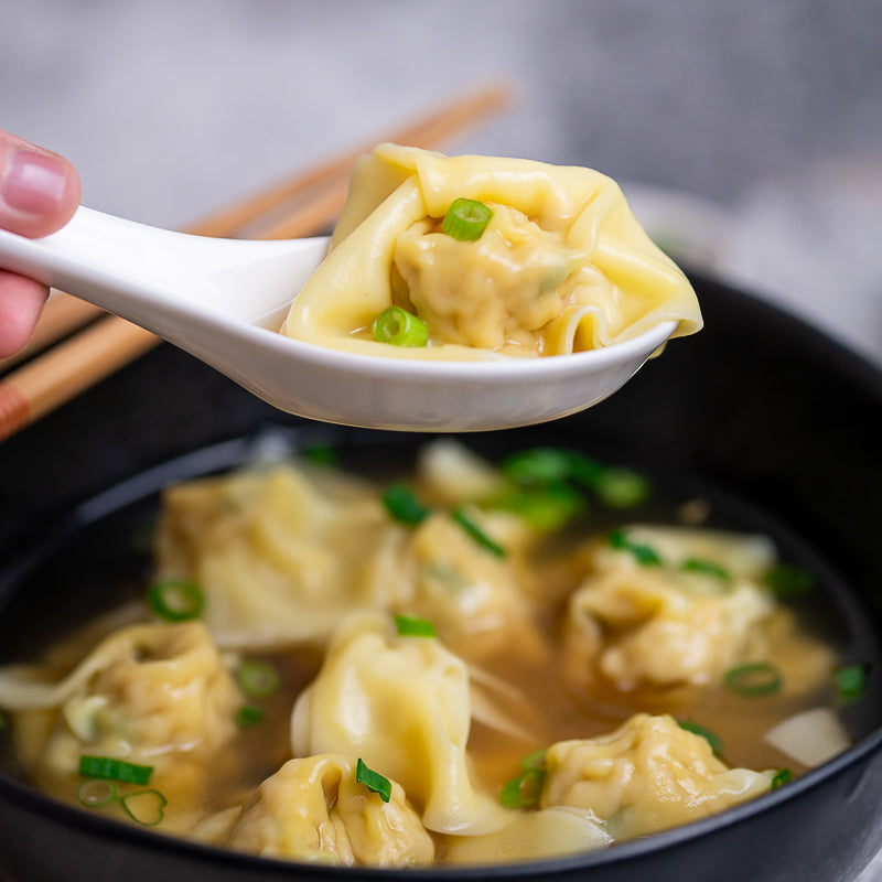 Spoon holding a wonton above a bowl of wonton soup with green onions.