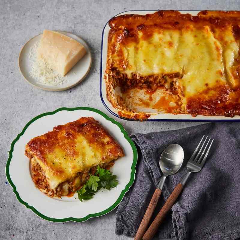 Baked lasagna with a slice on a plate, placed on a gray surface with a block of cheese and cutlery.